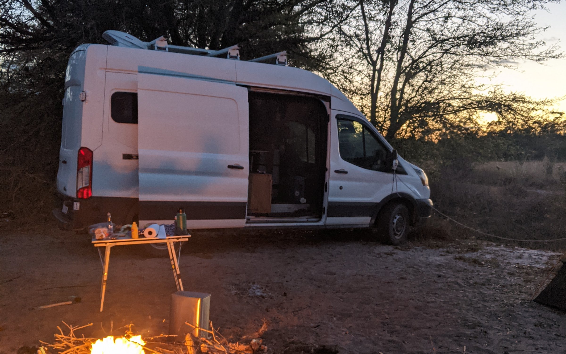 2017 Ford Transit T-250 high-roof campervan parked at dusk with the sliding door open and a campfire burning in the foreground