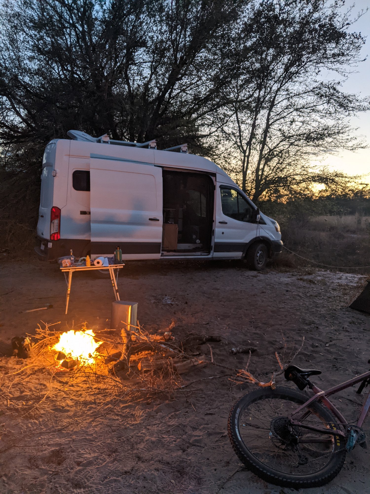 The van at a campsite at dusk, side door open, campfire burning in front